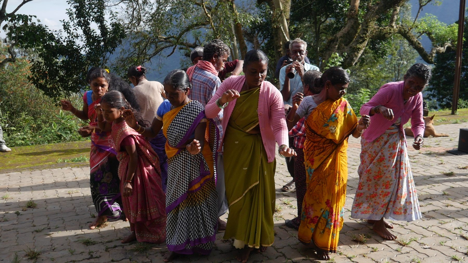 Women in colourful saris dancing together on a paved hilltop courtyard while a man plays a horn behind them